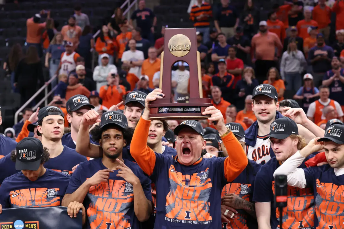 Mar 28, 2026; Houston, TX, USA; Illinois Fighting Illini head coach Brad Underwood and players celebrate with the trophy on the podium after defeating the Iowa Hawkeyes in an Elite Eight game of the South Regional of the men's 2026 NCAA Tournament at Toyota Center. Mandatory Credit: Troy Taormina-Imagn Images