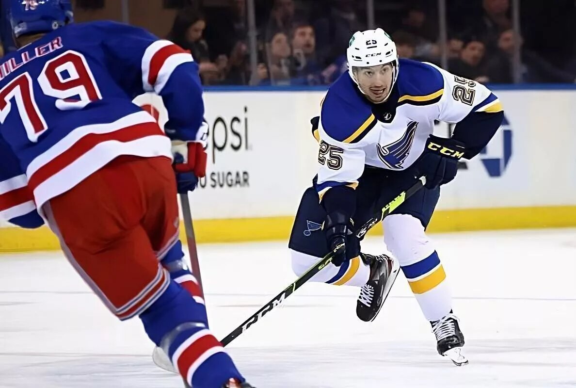 St Louis Blues winger Jordan Kyrou (#25, white jersey), skates with the puck as New York Rangers defenseman K'Andre Miller (#79, blue jersey) defends.