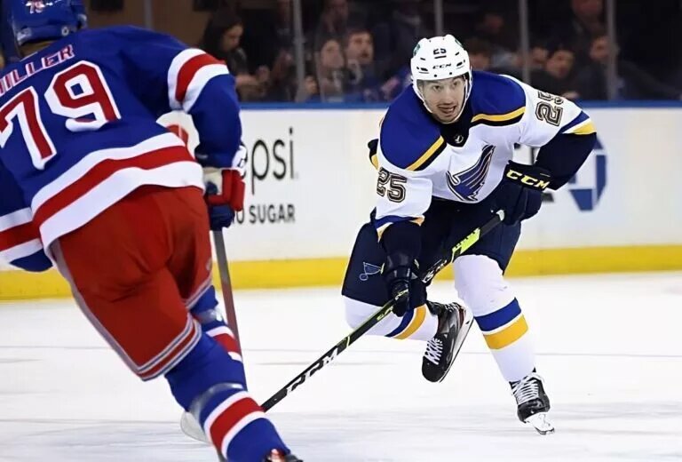 St Louis Blues winger Jordan Kyrou (#25, white jersey), skates with the puck as New York Rangers defenseman K'Andre Miller (#79, blue jersey) defends.
